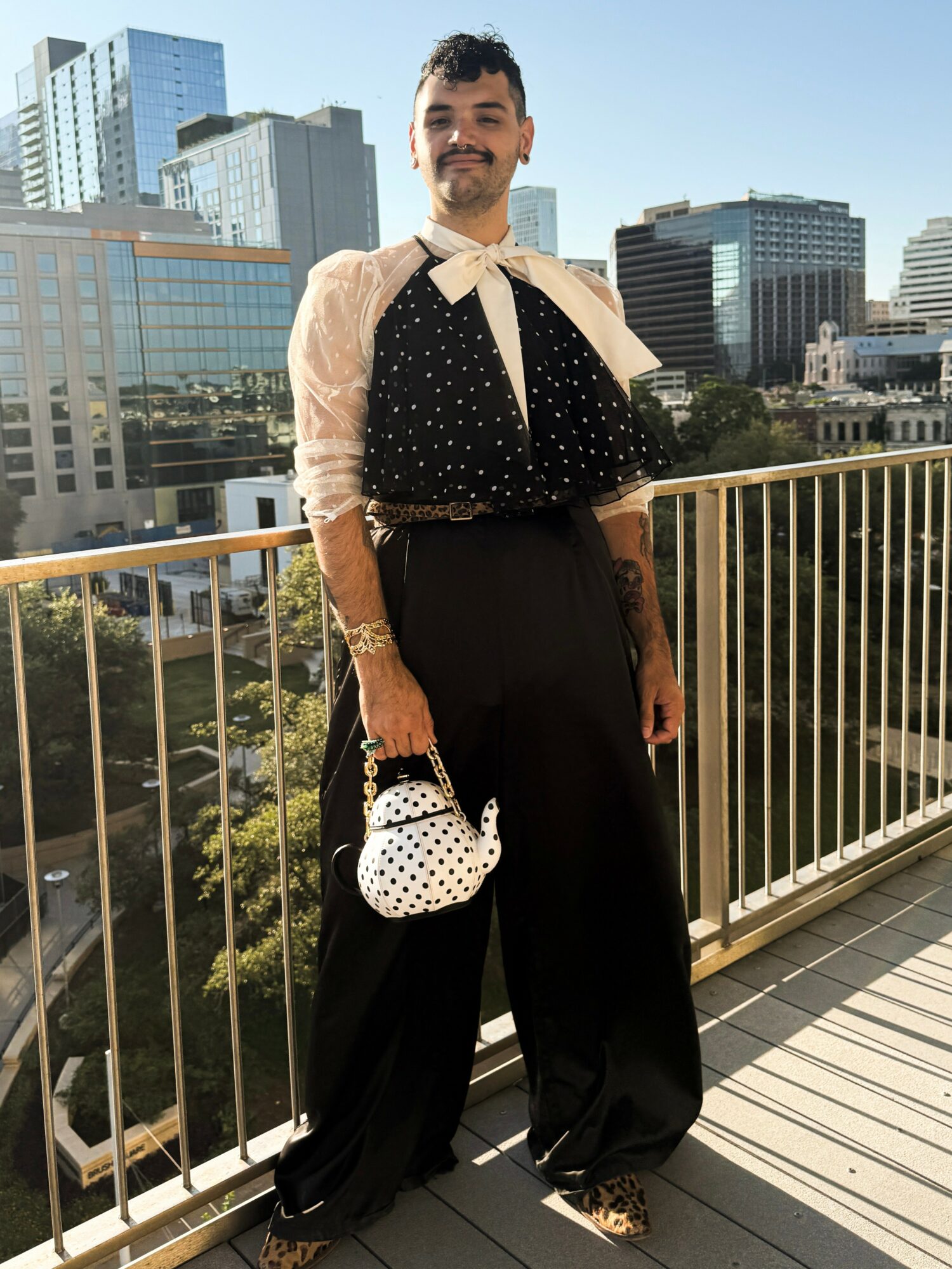 Person standing on balcony with city skyline in background, wearing a polka dot top, white bow, and black pants, holding a polka dot bag.