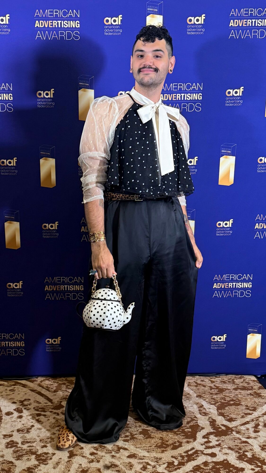 Person standing on patterned carpet in front of blue backdrop with logos, wearing a black and white outfit, holding a polka dot bag.