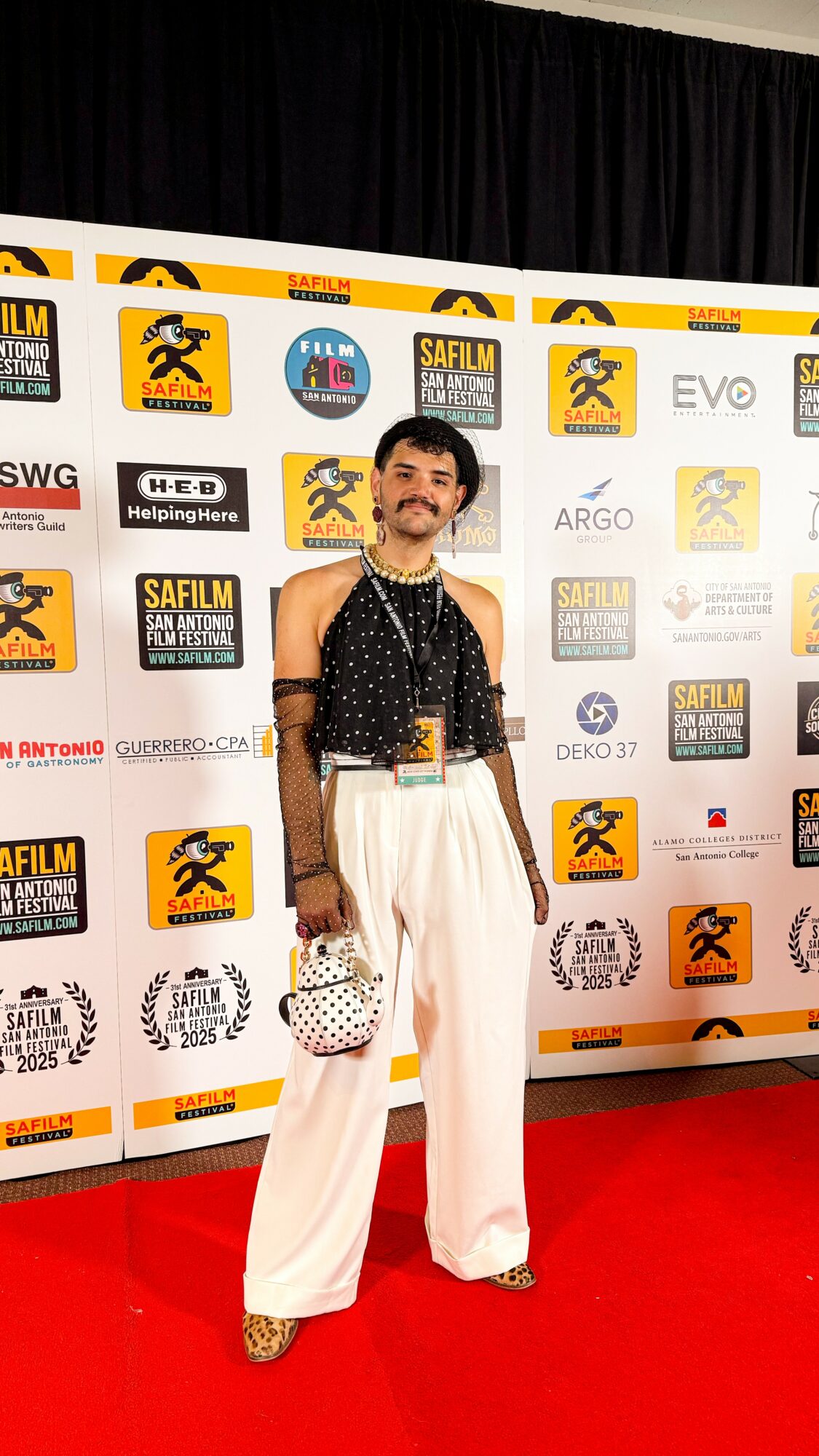 Person standing on red carpet in front of a backdrop with logos, wearing a sleeveless top and white wide-leg pants, holding a small bag.