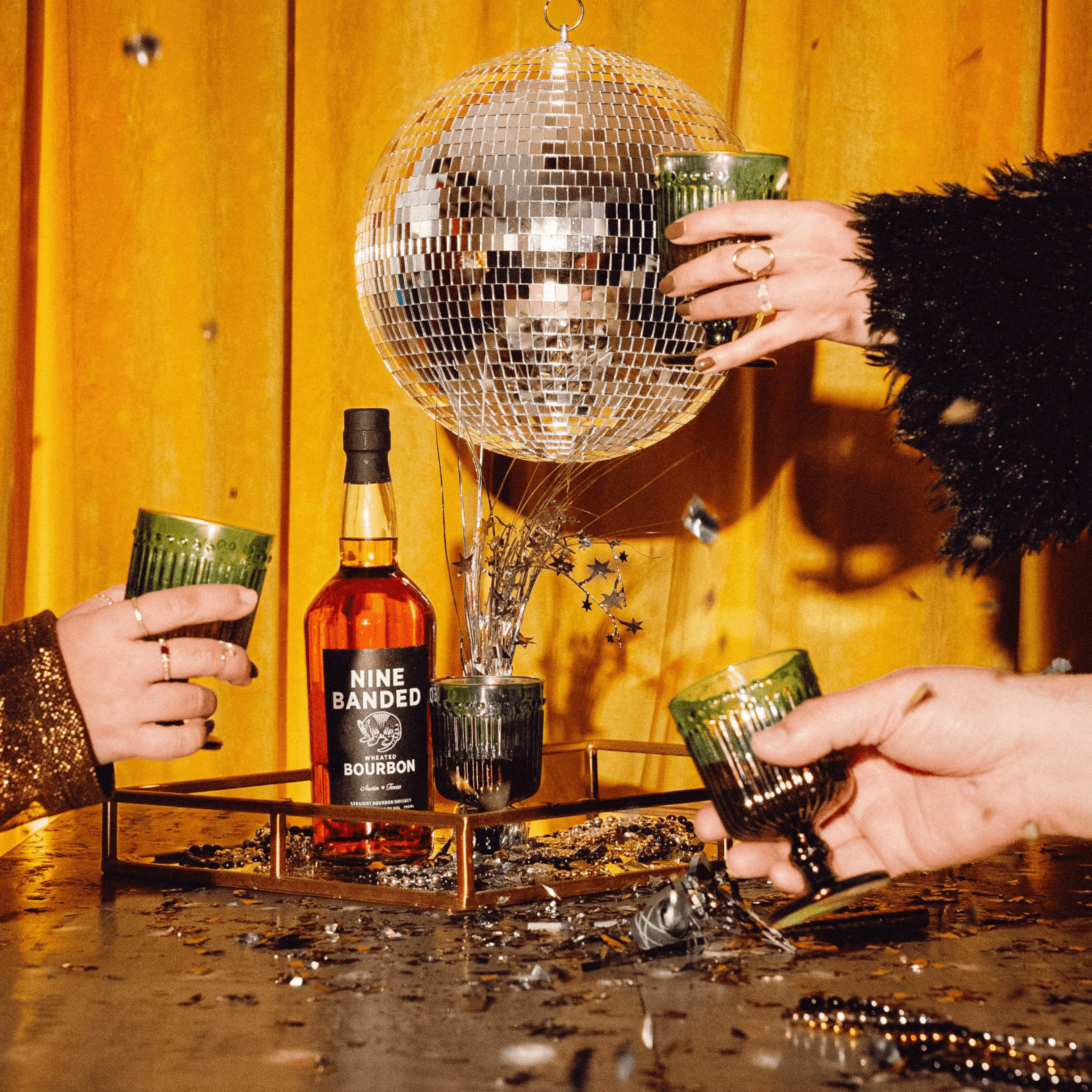 People's hands holding glasses around a bottle, with a disco ball and water splash on a table, yellow curtain background.