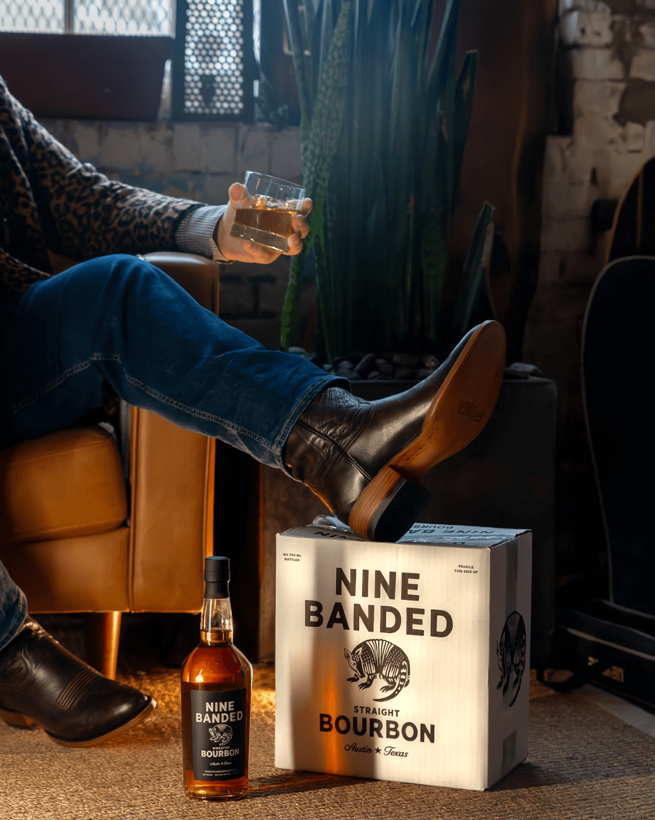 Person sitting on a sofa holding a glass of drink, with a whiskey bottle and box of Nine Banded Bourbon nearby, in a cozy setting.