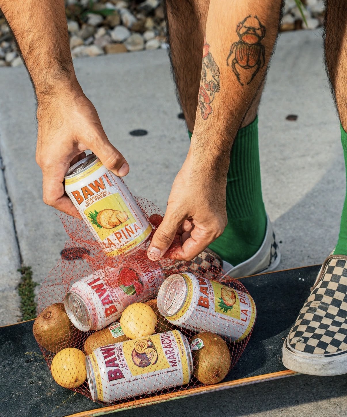 Person's hand holding a can of BAWL La Pina, with cans and tennis balls on the ground, and legs in green socks and checkered shoes.