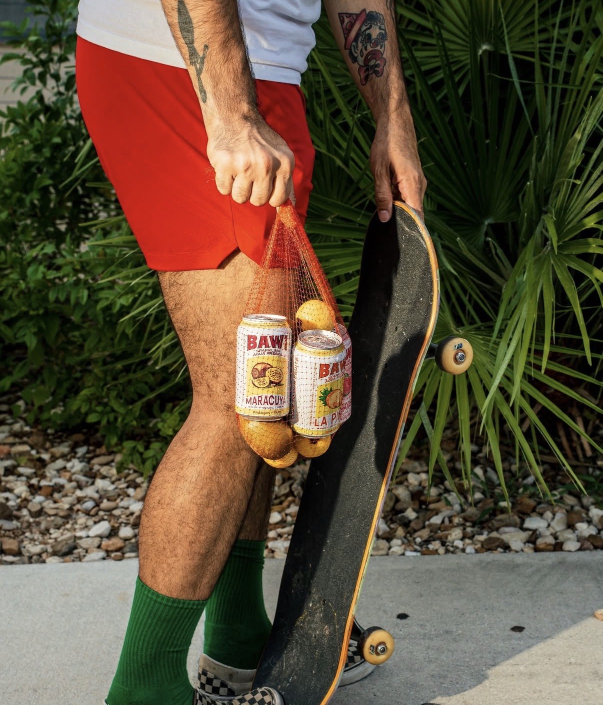 Person holding a skateboard and a bag with cans and a ball, wearing red shorts and green socks, outdoors.