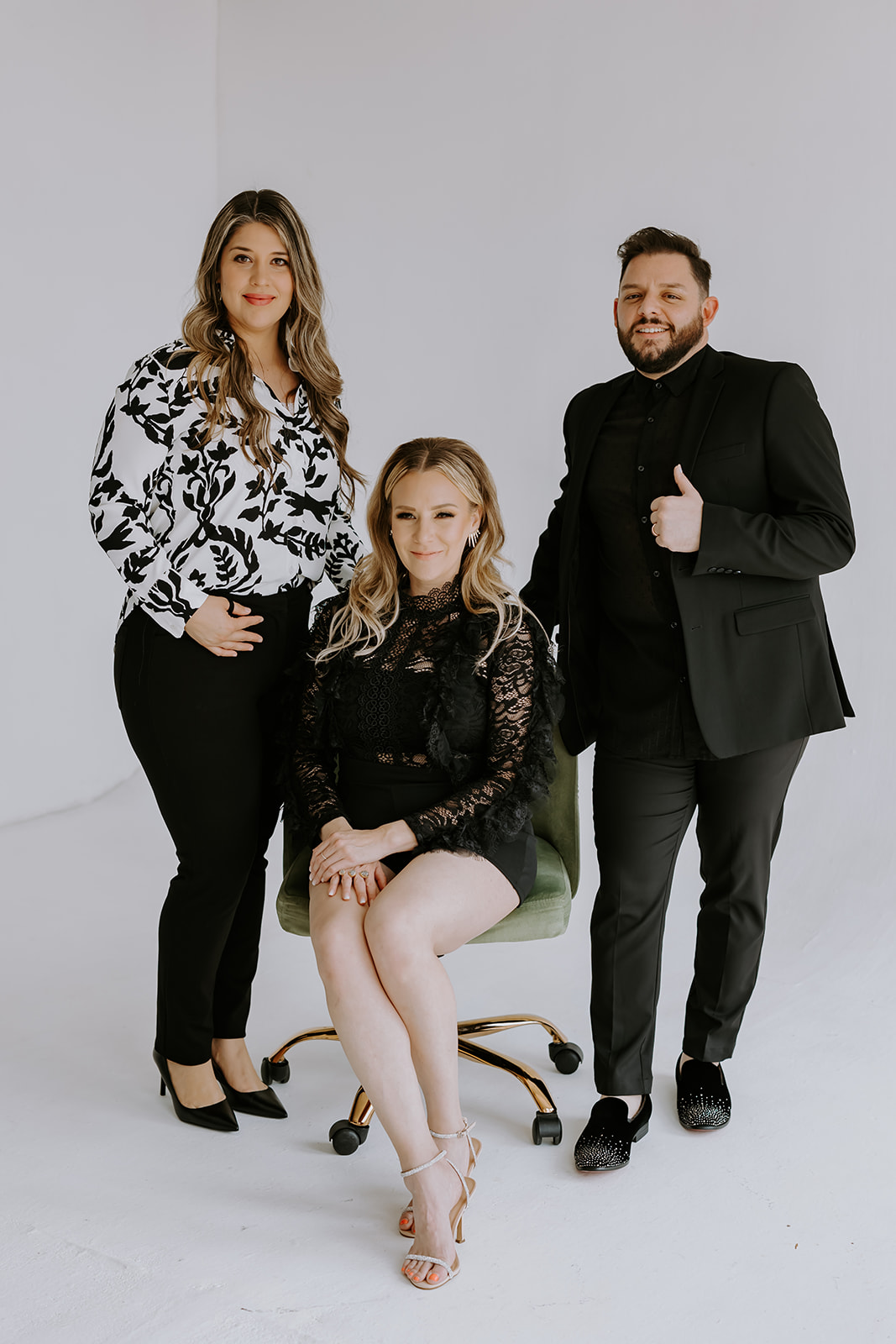 Three people pose together in a studio with a plain background; two standing and one sitting on a chair.