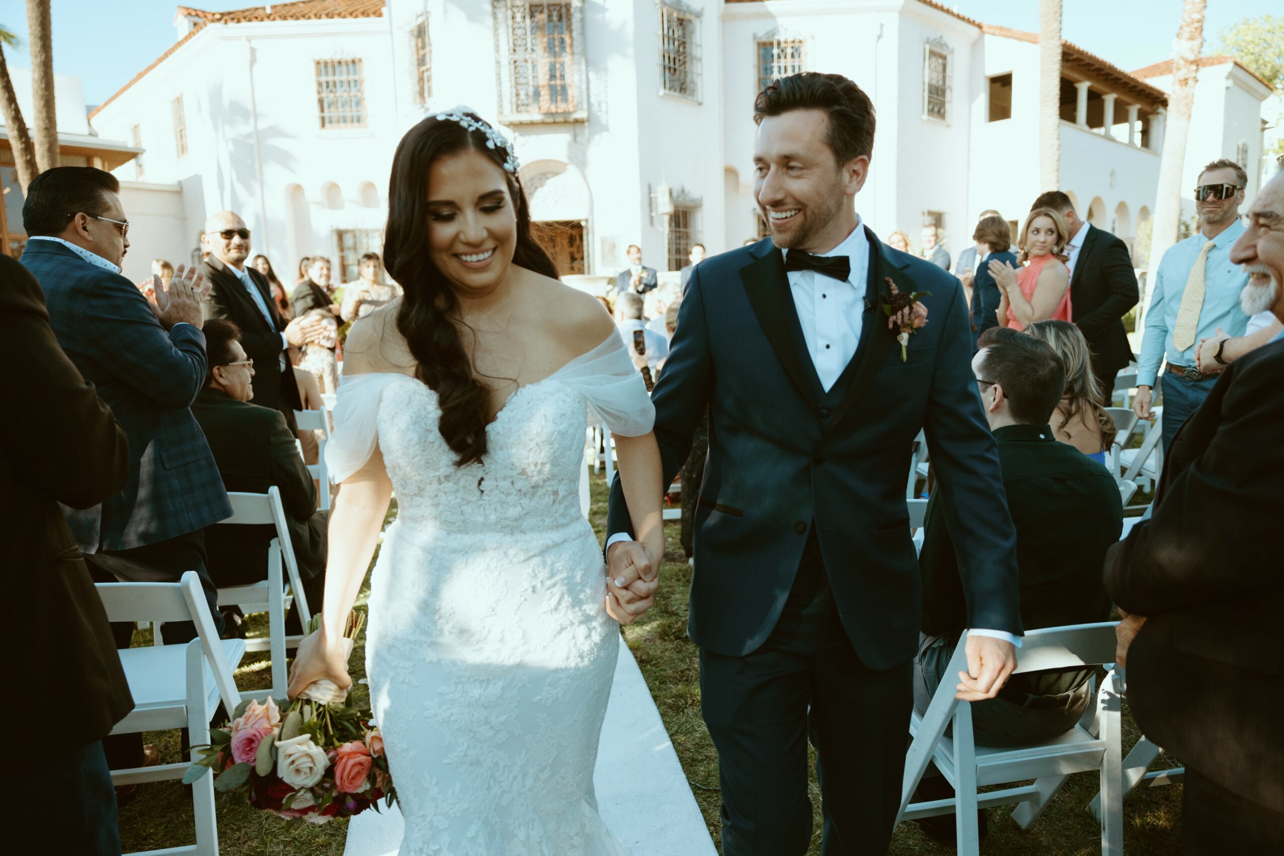 Bride and groom holding hands, smiling, outdoors wedding ceremony with seated guests and white buildings in background.