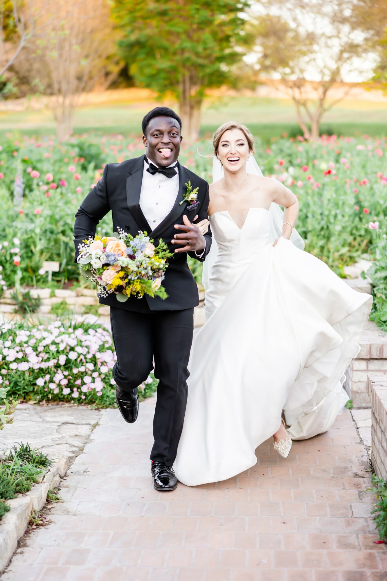 A joyful couple in wedding attire walking outdoors on a brick path, surrounded by colorful flowers and greenery.
