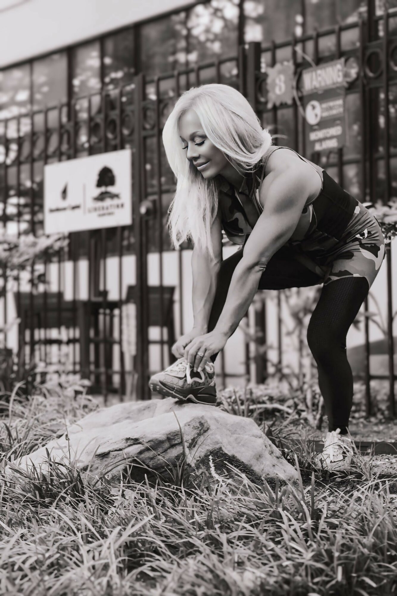 Woman with long hair bending down to pick up a rock outdoors, with a fence and signs in the background.