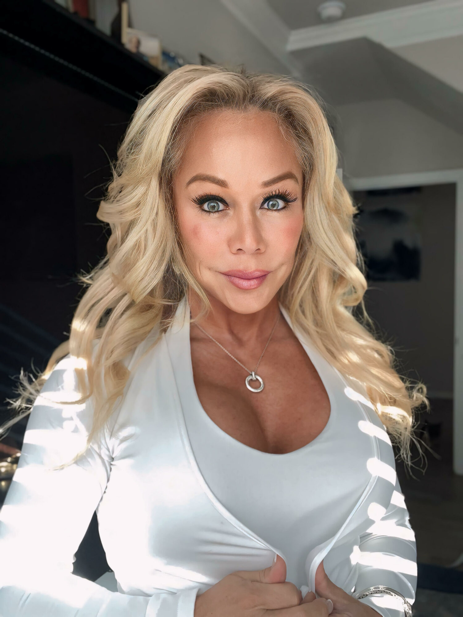 Woman with long blonde hair and blue eyes taking a selfie indoors, wearing a white top and a necklace with a heart pendant.