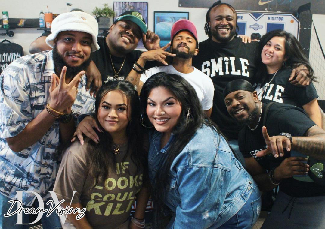 Group of nine diverse people smiling and posing together indoors, some making hand gestures, with a background of posters and equipment.