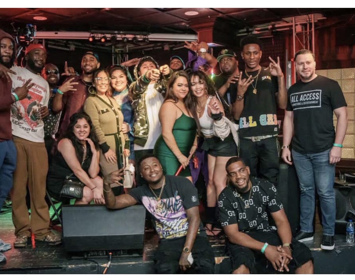 Group of people posing together in a club or event space, some making hand gestures, smiling, and dressed casually.