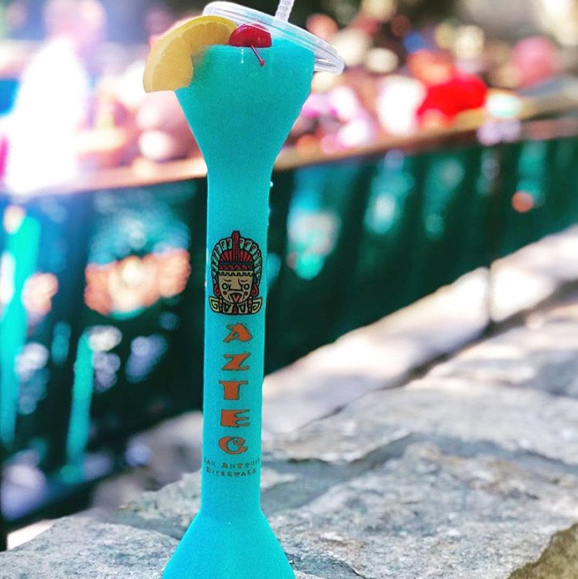 Blue frozen drink with lemon wedge and cherry, on a table outdoors, blurred people in background.