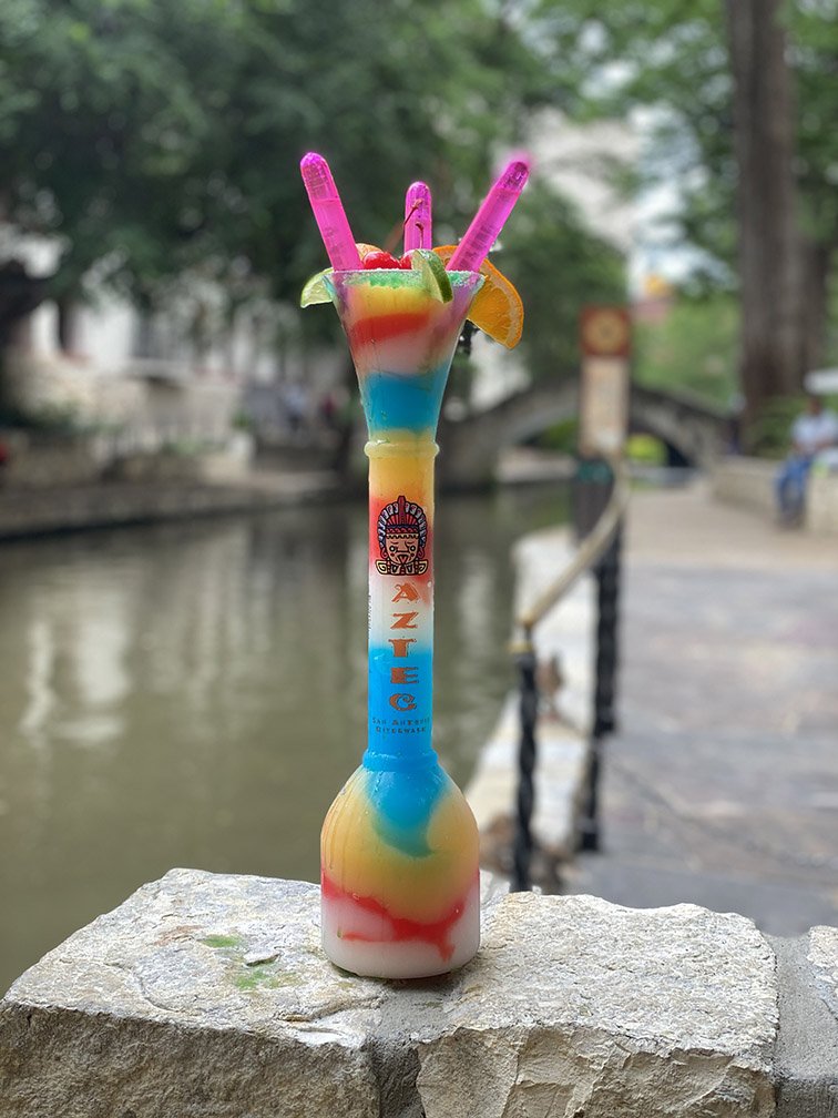 Colorful rainbow layered drink with pink straws and orange slices on a stone surface outdoors.