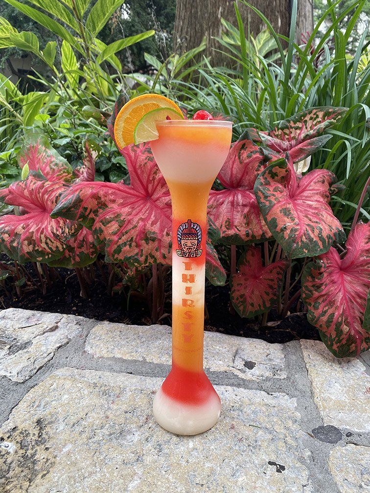 Colorful cocktail in a tall glass with a lemon slice garnish, set outdoors among green plants and pink leaves.