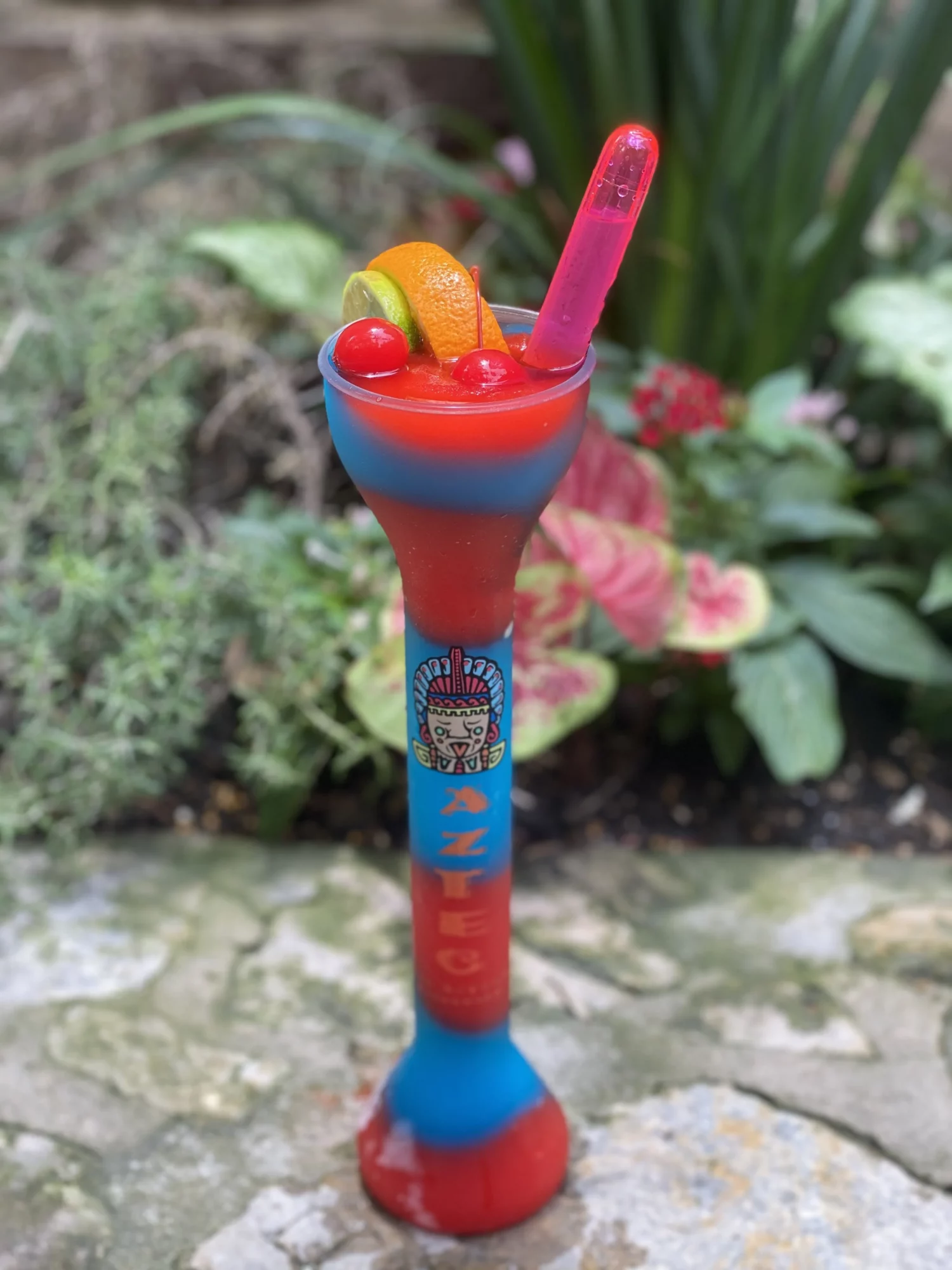 Colorful cocktail glass with fruit garnishes and a pink straw, set outdoors on a stone surface with plants in background.