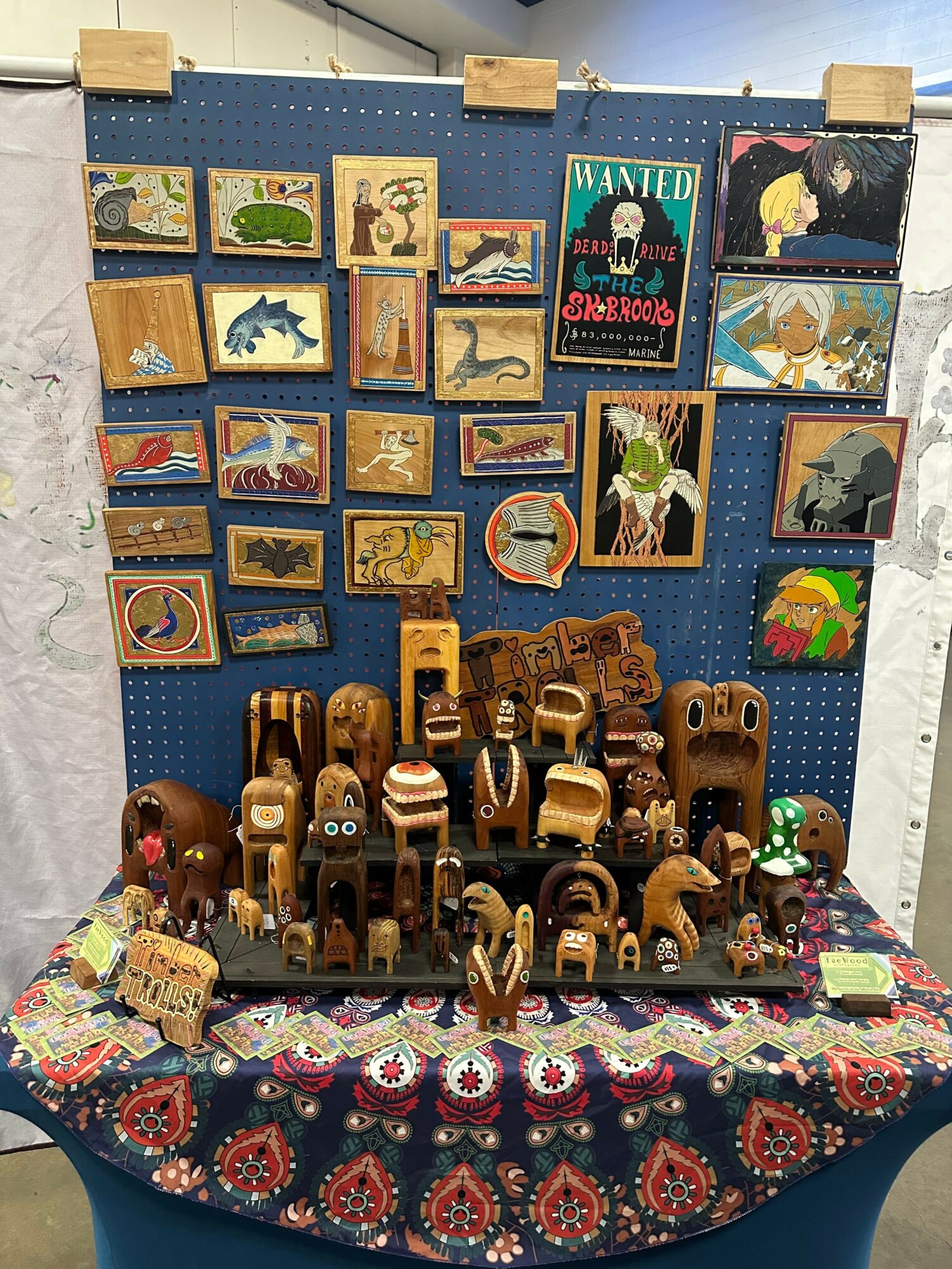 Display of small paintings and wooden carved figurines on a table with a colorful cloth, against a blue pegboard background.