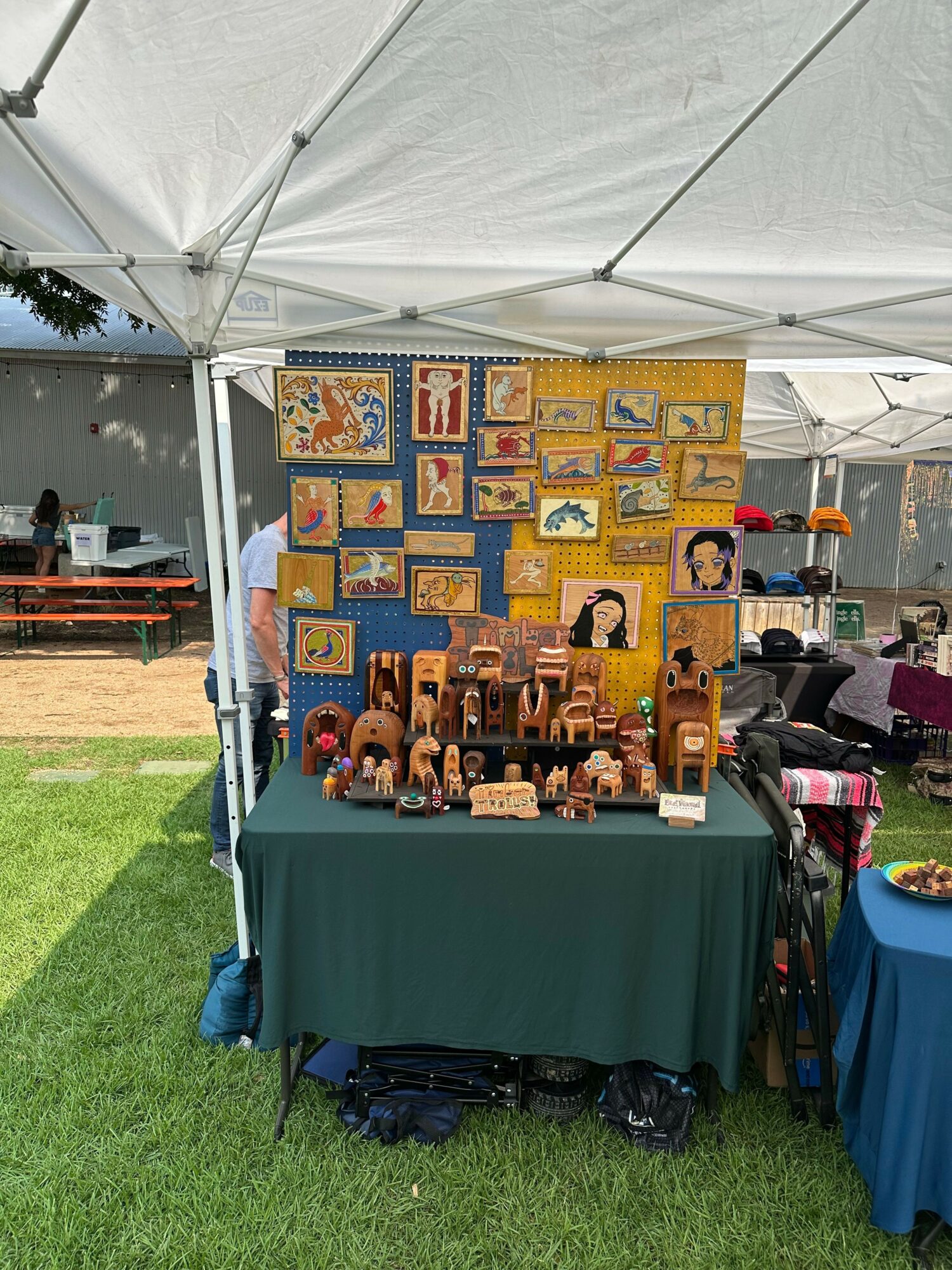 Display of colorful quilted artwork and framed pictures on a yellow wall, with a table covered in a green cloth and bags on the grass.