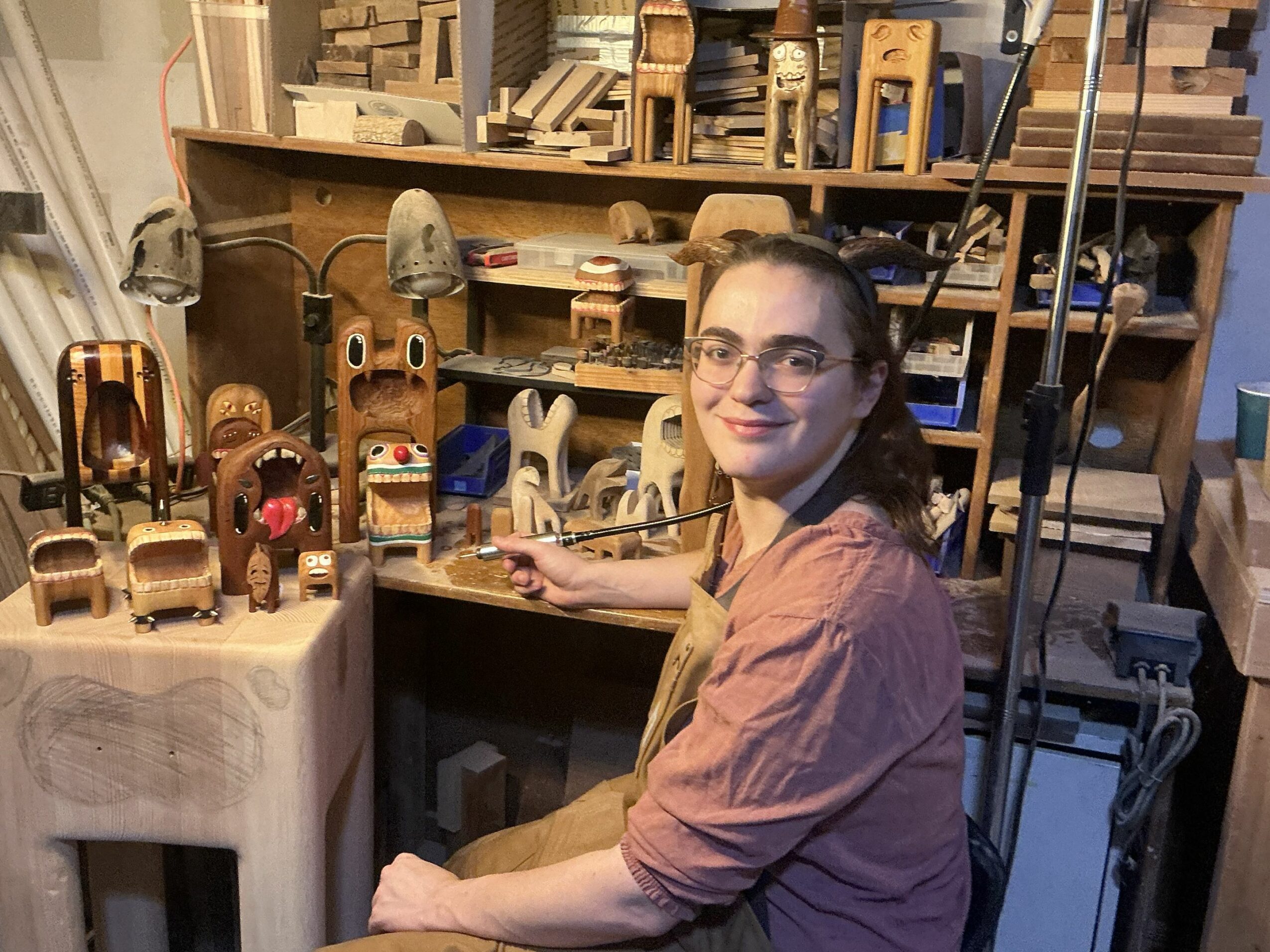 Young woman with glasses and a pink shirt sitting in a woodworking shop surrounded by wooden crafts and tools.