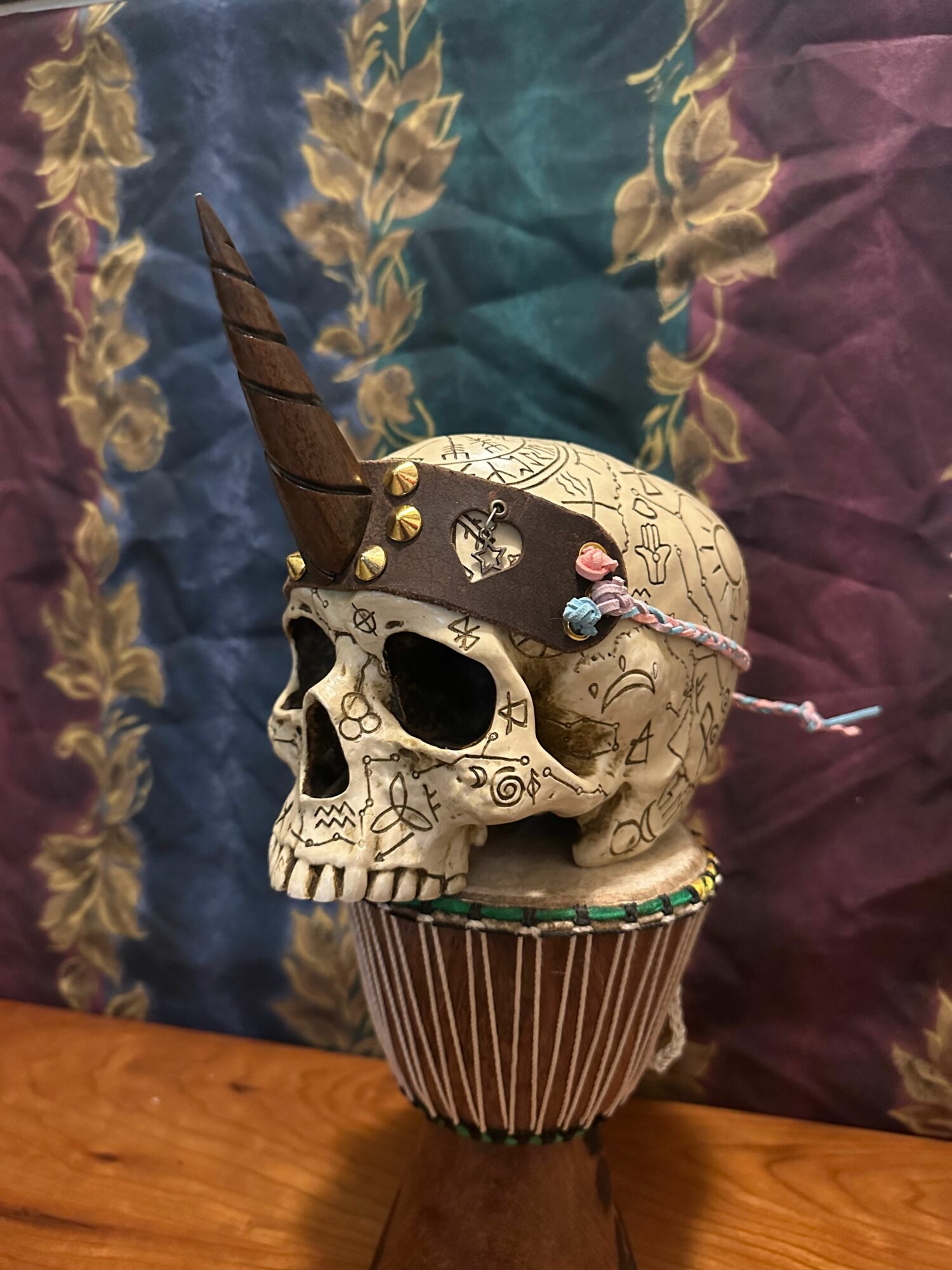 Decorative skull with a candle on top, surrounded by a feathered headdress, on a reflective surface with a floral pattern.