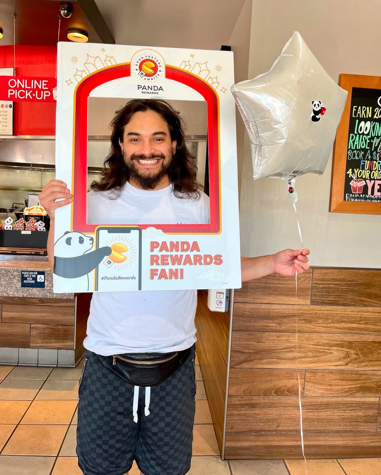 Person holding a panda-themed photo frame with a smiling face, holding a star-shaped balloon, in a store or cafe setting.