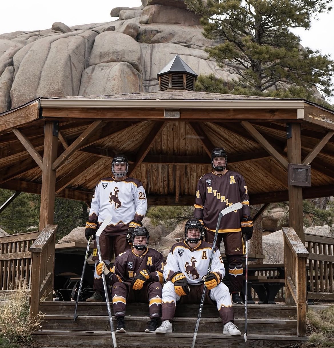Four hockey players in uniforms sitting and standing on a wooden pavilion with rocks and trees in background.