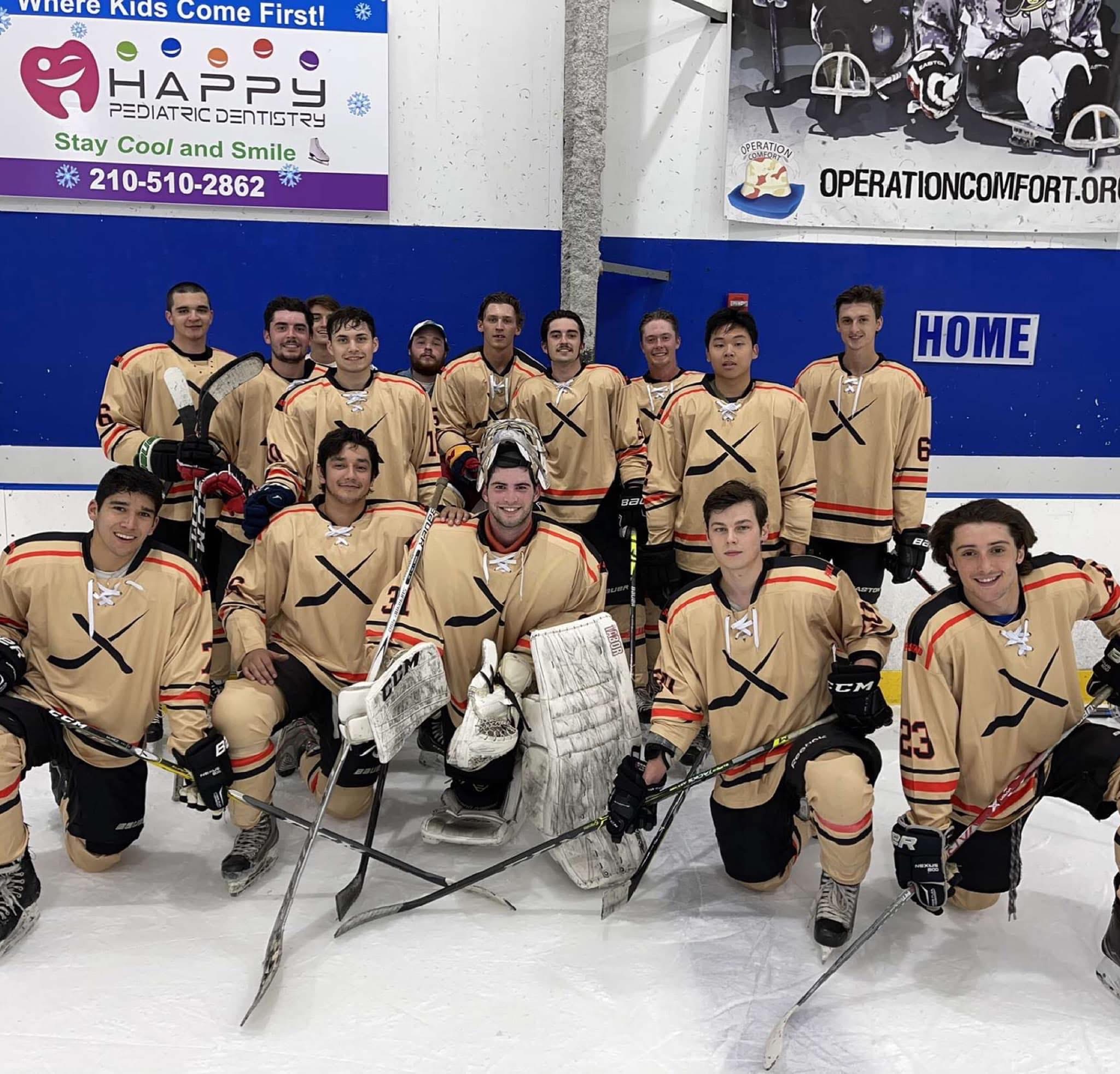 Group of young hockey players in beige jerseys on ice rink, some kneeling, some standing, with goalie in front center.