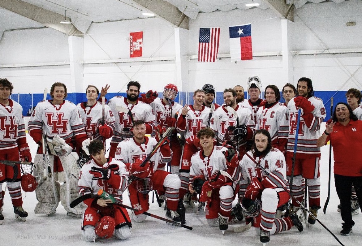 Group of ice hockey players in uniforms on ice rink, flags hanging above, team members posing for photo.