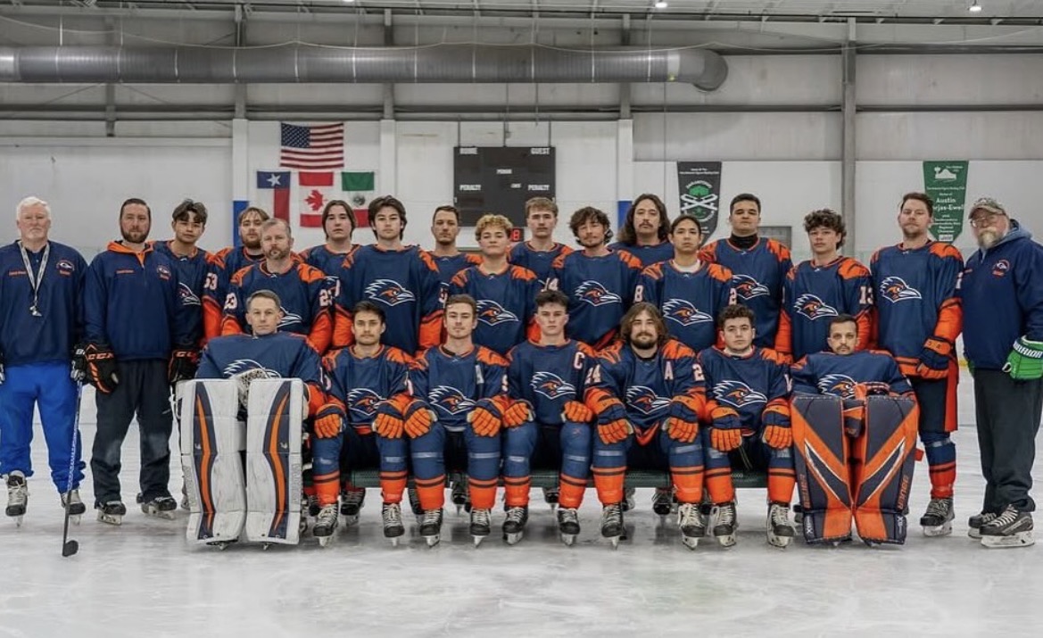Group of ice hockey players and coaches in team uniforms on ice rink, with flags in background.
