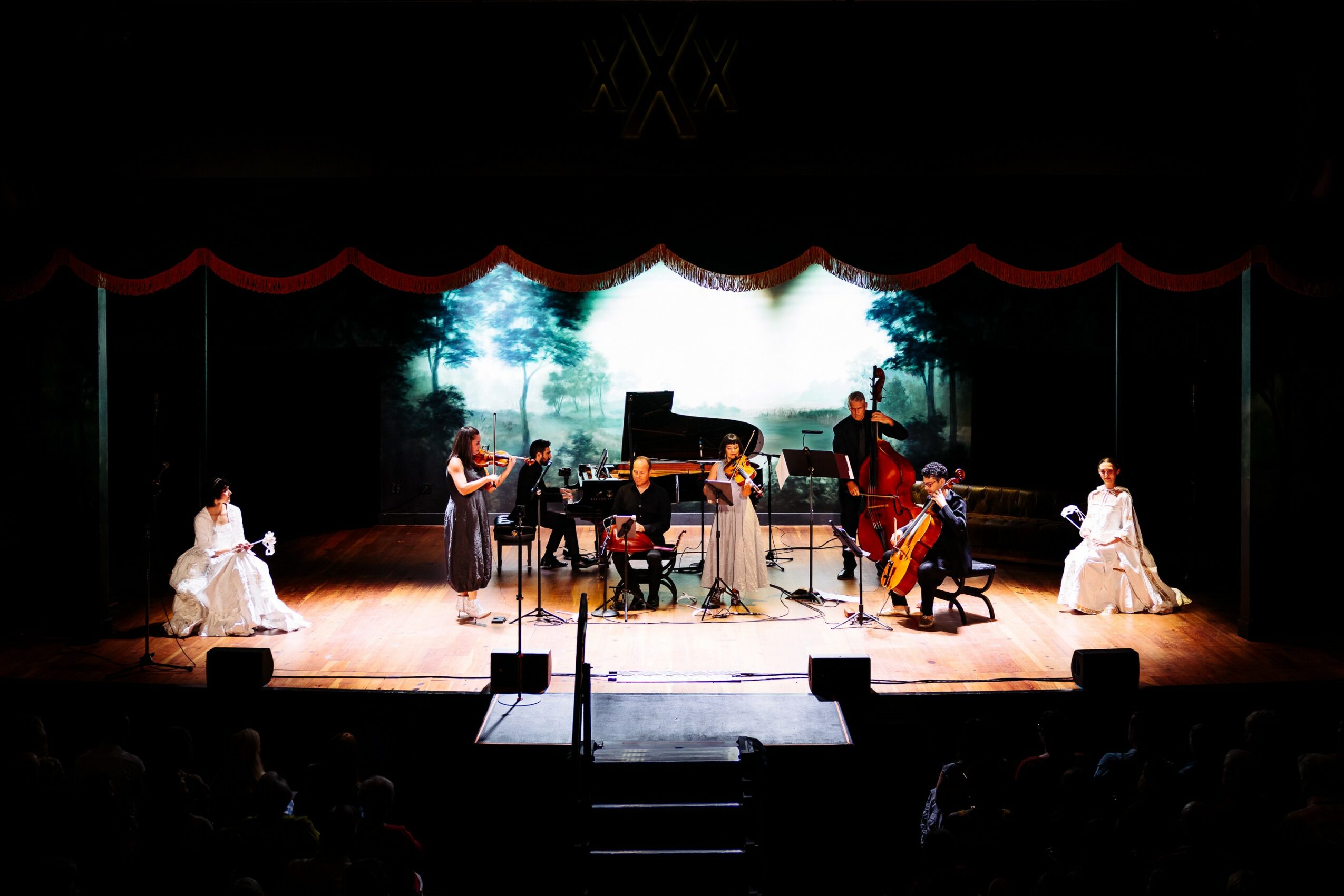 Musicians perform on stage with a backdrop of trees, two women in white dresses sit at sides, stage lit brightly.