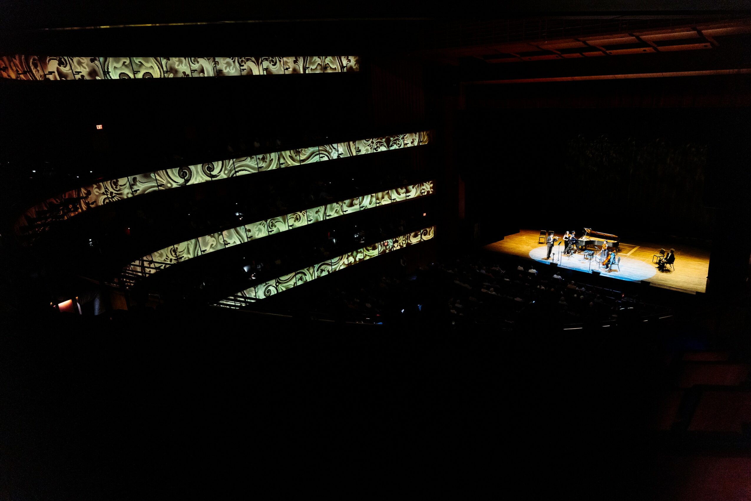 Dark theater with illuminated stage and patterned balcony, no visible audience, stage lights on, intricate balcony design.