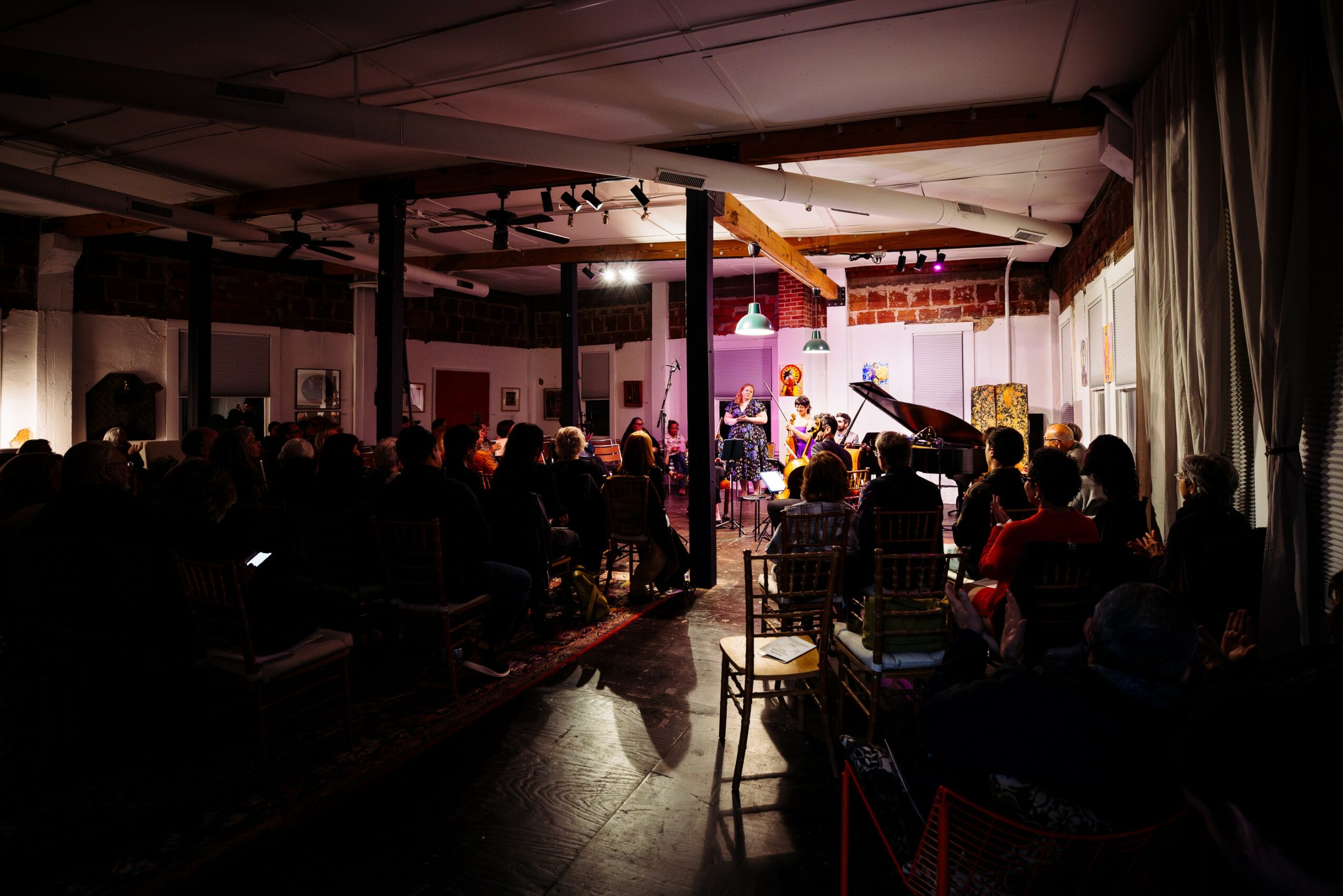 Stage with musicians performing, audience seated in a dimly lit room, curtains on the side, some light on stage.