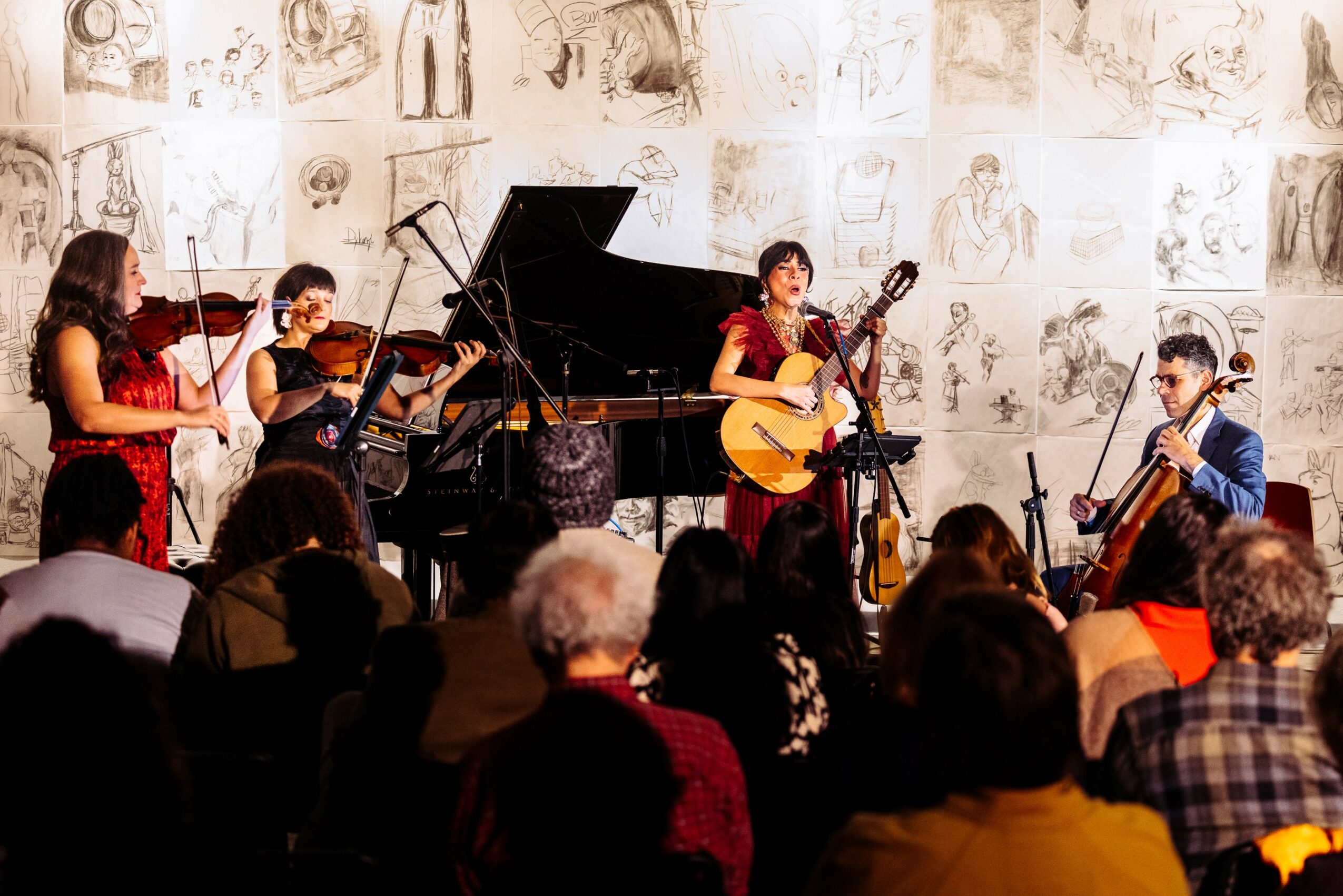 Group of five musicians performing on stage with a grand piano, audience watching, wall with sketches in background.