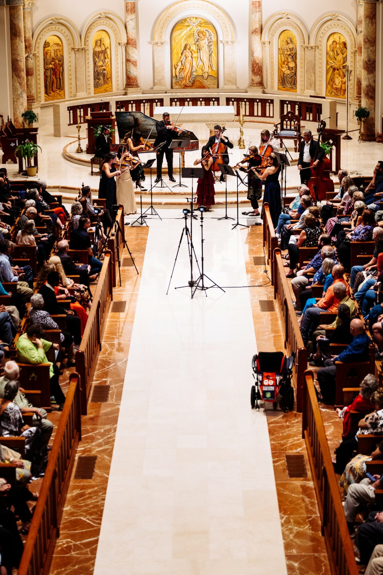 Orchestra performs in a church with an audience seated on both sides, ornate gold decorations, and a grand altar in the background.