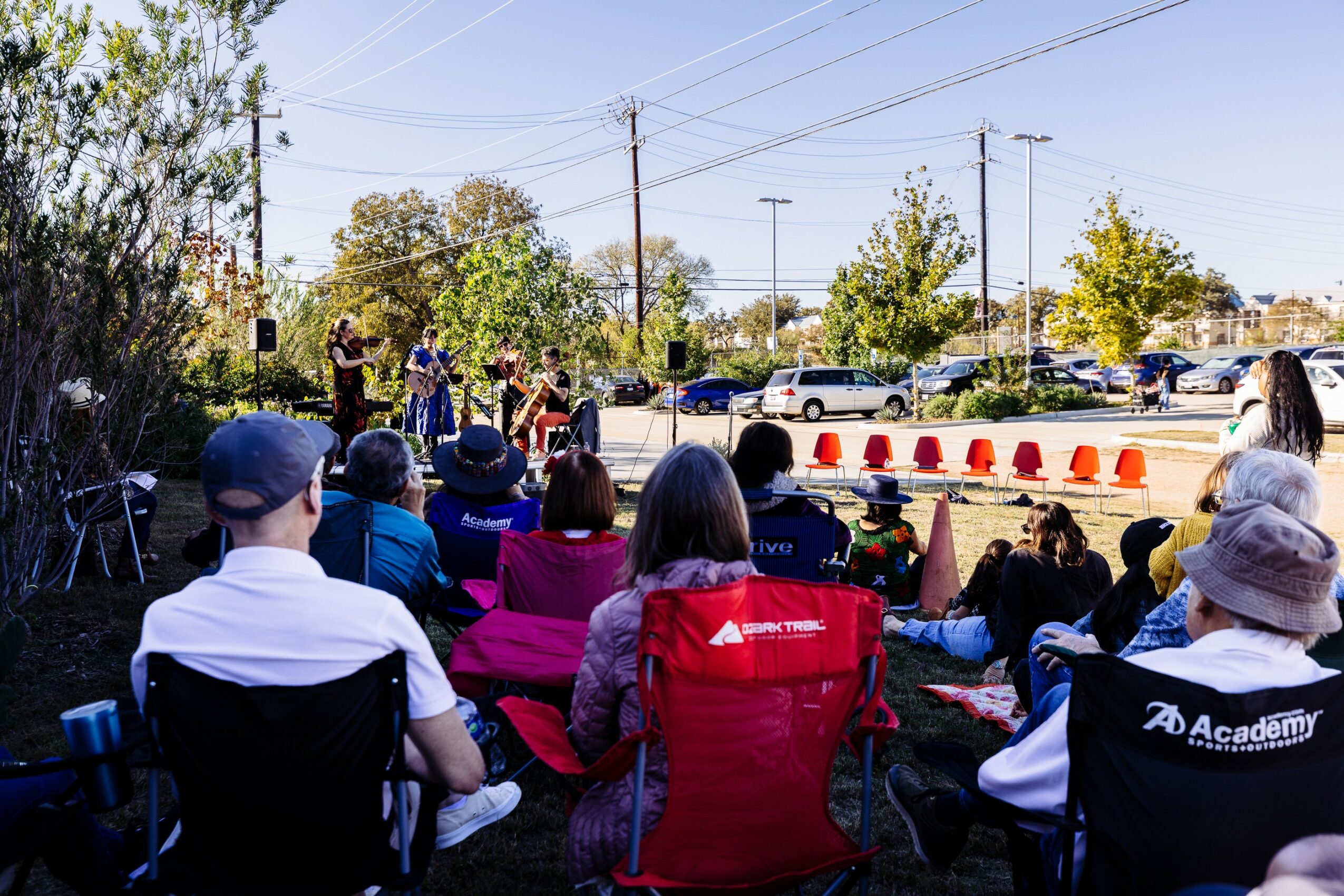 Group of people seated outdoors watching a performance on a sunny day, with trees and parked cars in background.