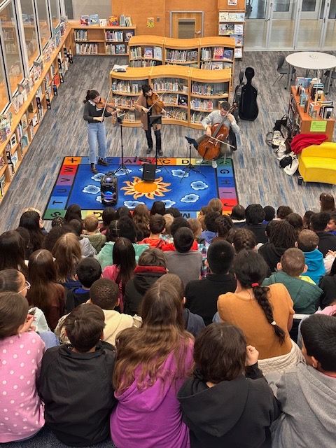 Children gathered in a library listening to a person reading a book, with bookshelves in the background.