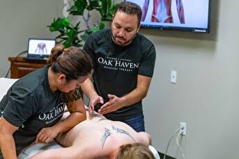 Two people perform a medical procedure on a person's back in a room with a TV and plant, one person is lying down.