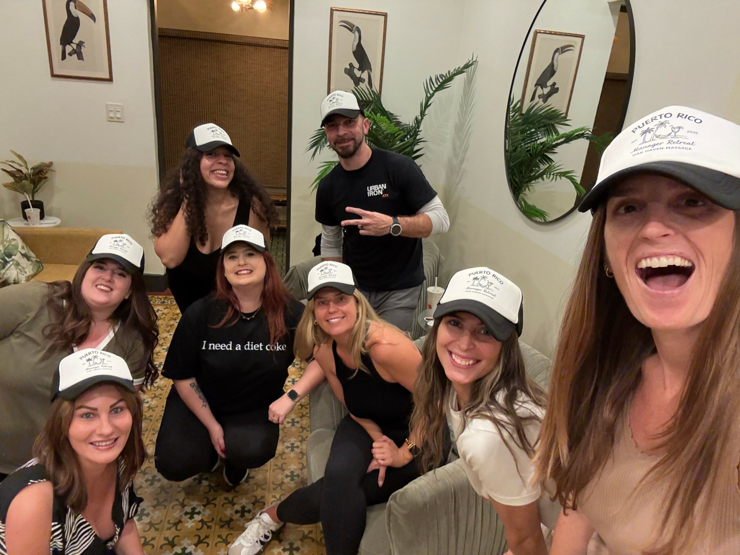 Group of seven people wearing white caps, smiling indoors with plants and framed art, taking a selfie.