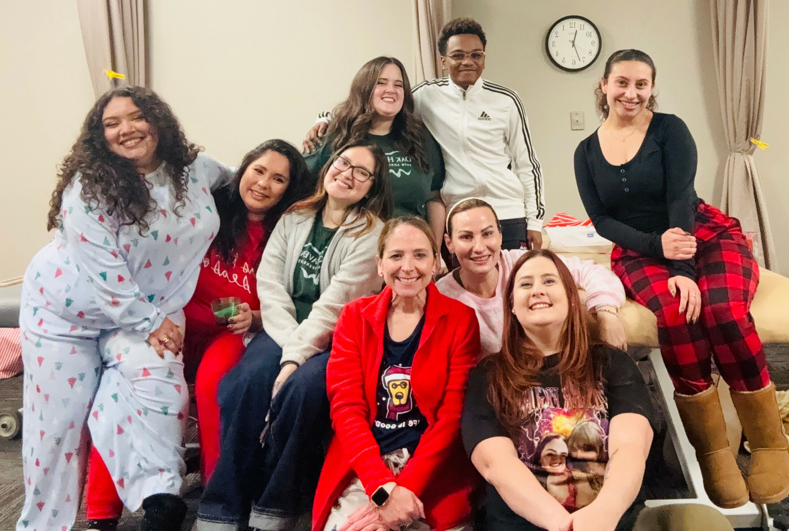 Group of ten people smiling indoors, some sitting and some standing, in casual and festive clothing, with a clock on the wall.