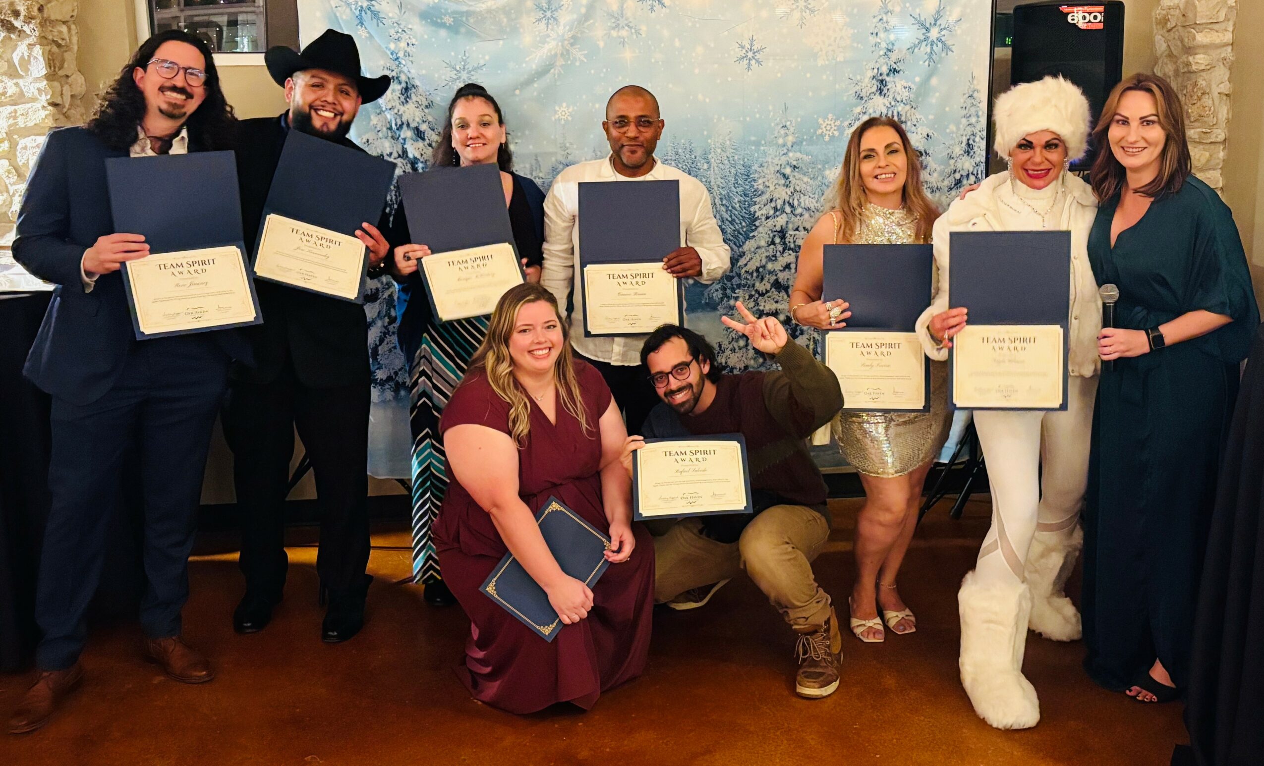 Group of ten people holding certificates, some wearing festive attire, in front of a winter-themed backdrop.