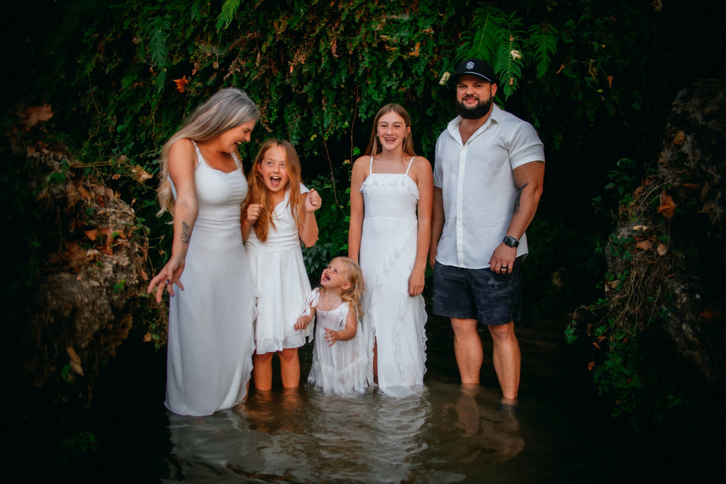 Group of six people standing in water outdoors, surrounded by greenery, smiling, with four women and two children, one man.