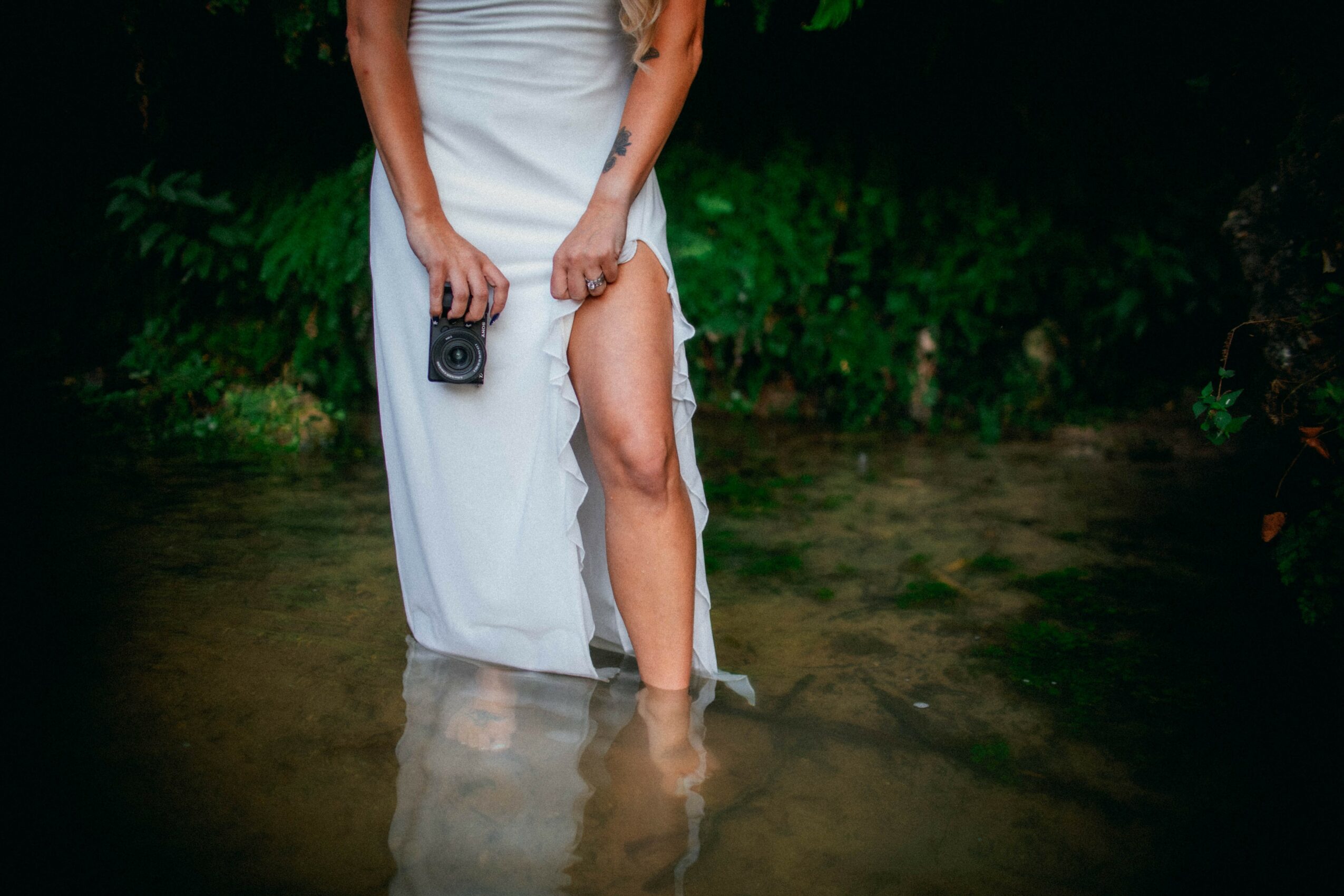 Person standing in water holding a camera, wearing a long dress with a slit, surrounded by greenery.