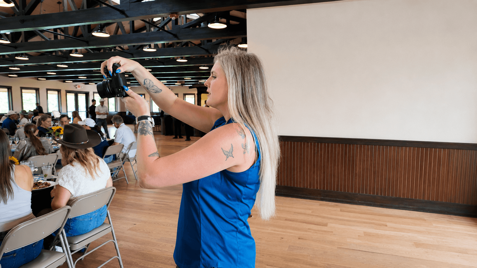 Woman with long blonde hair wearing a blue sleeveless top holding a camera in a large indoor space with people seated at tables.