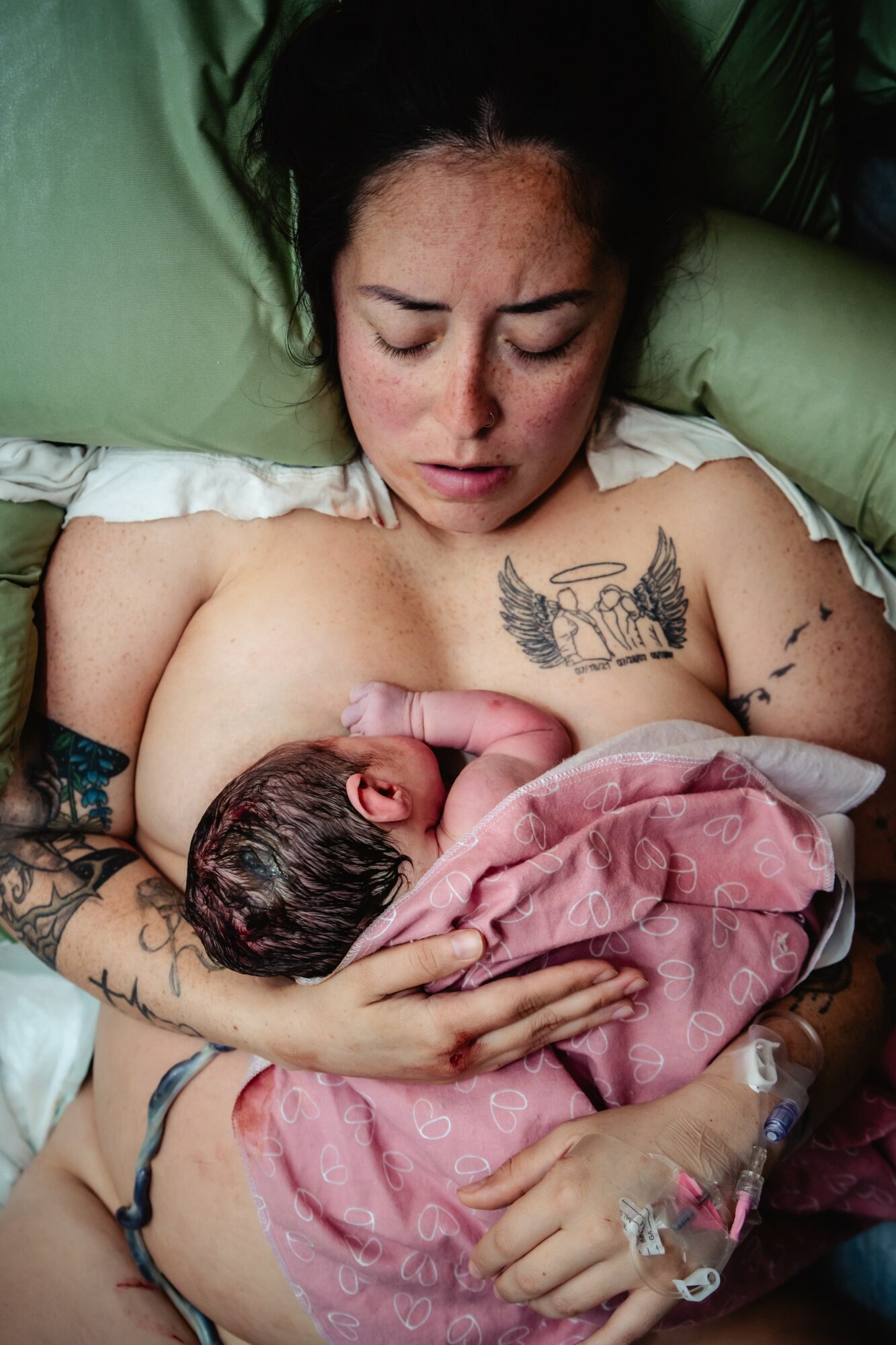 Woman lying in bed holding a newborn baby wrapped in a pink blanket, with a tattoo on her chest.