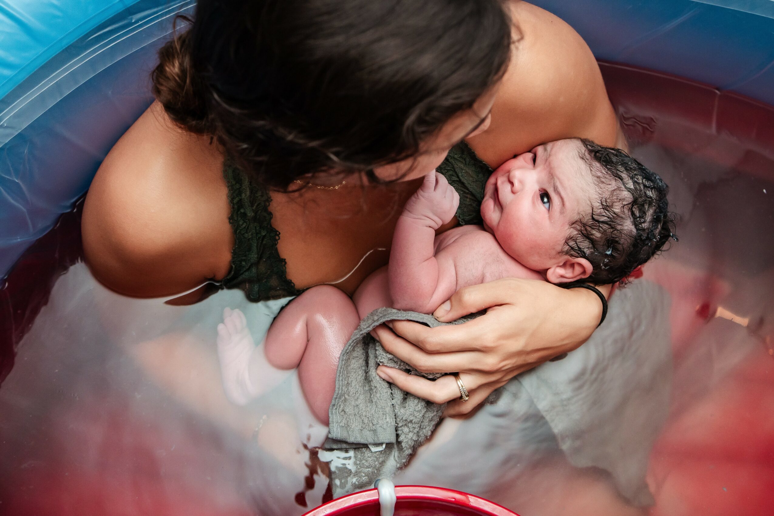 Woman holding a newborn baby in a small pool, water around them, woman looking at baby, baby wrapped in cloth