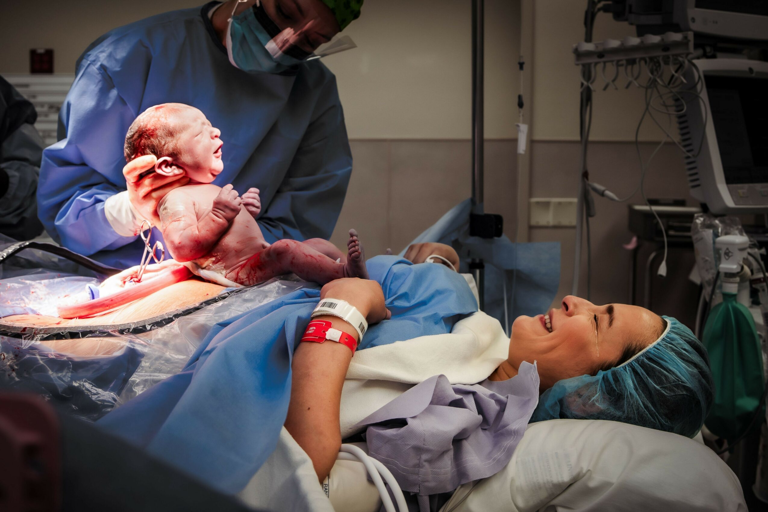 A newborn baby is held by a healthcare worker in a hospital room, with a woman lying on a bed nearby. Medical equipment is visible.