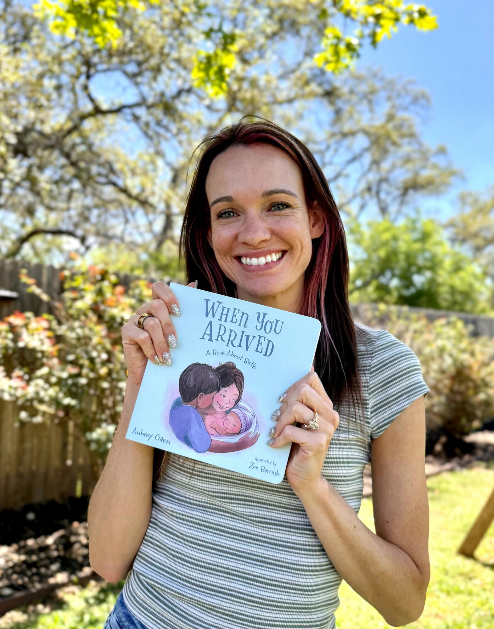 Woman holding a book outdoors, smiling, with trees and blue sky in background.