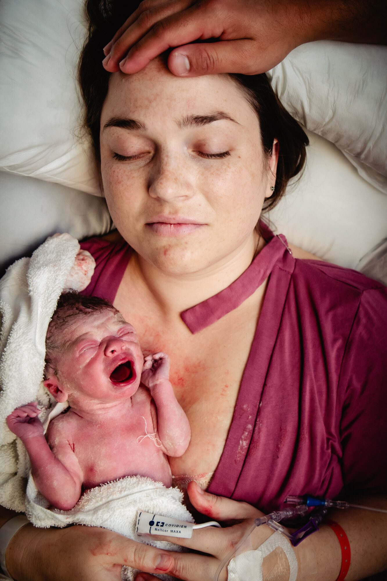 Woman lying down with eyes closed, holding a crying newborn baby on her chest, both on a bed.