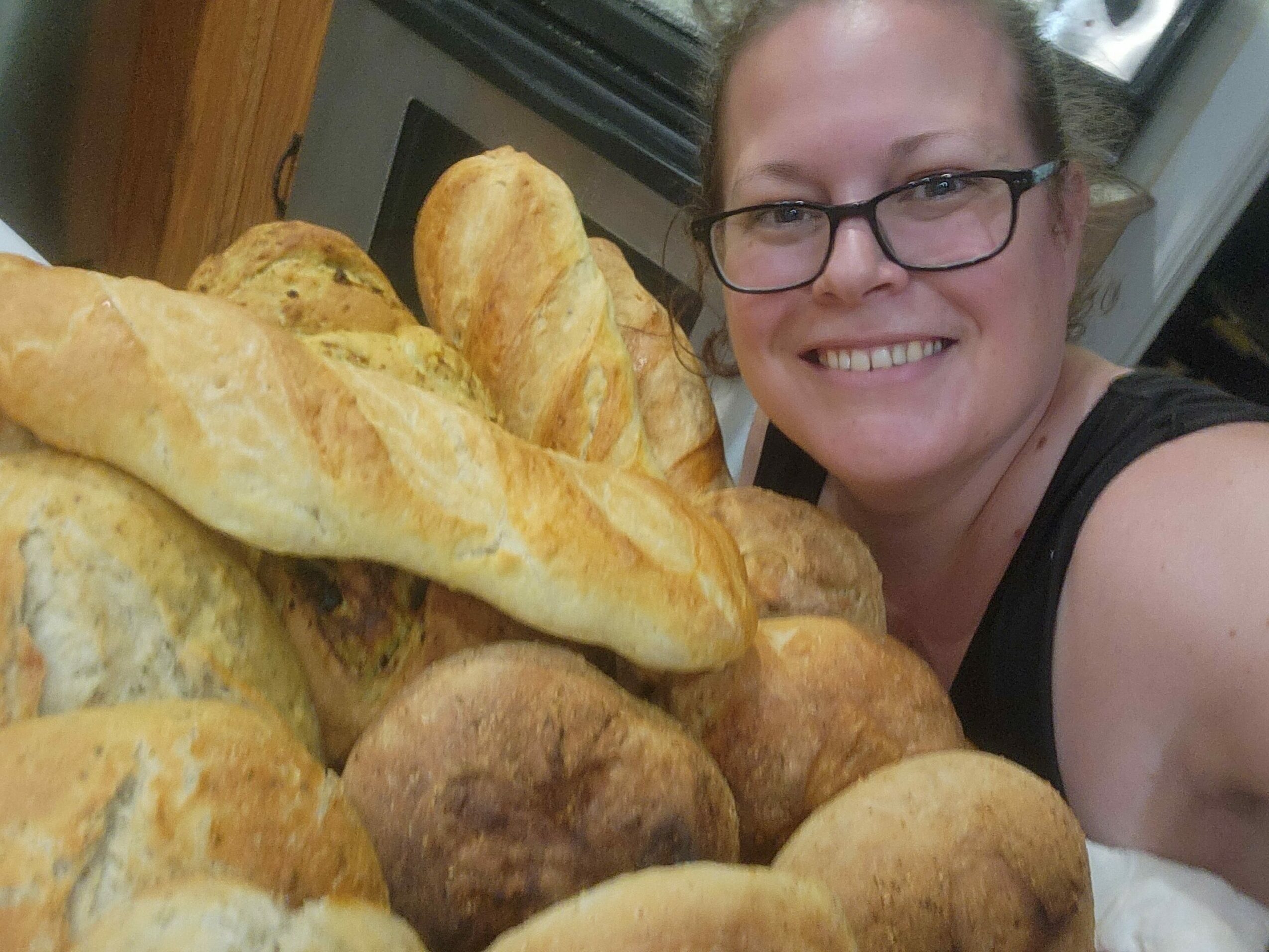 Person smiling with glasses, surrounded by large bread loaves, in a kitchen setting.