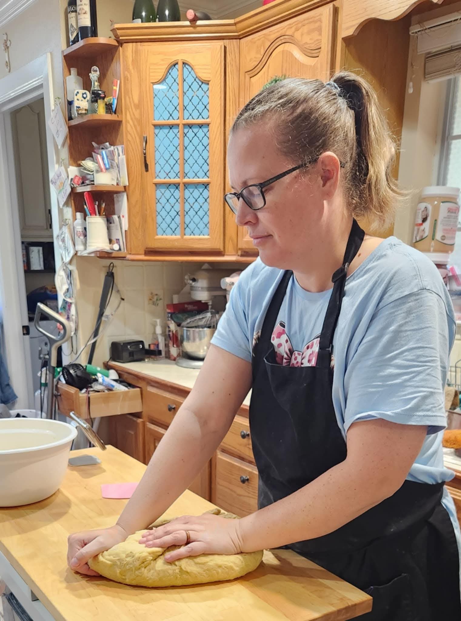 Woman wearing glasses and apron rolling dough on a kitchen counter, with wooden cabinets and shelves in background.
