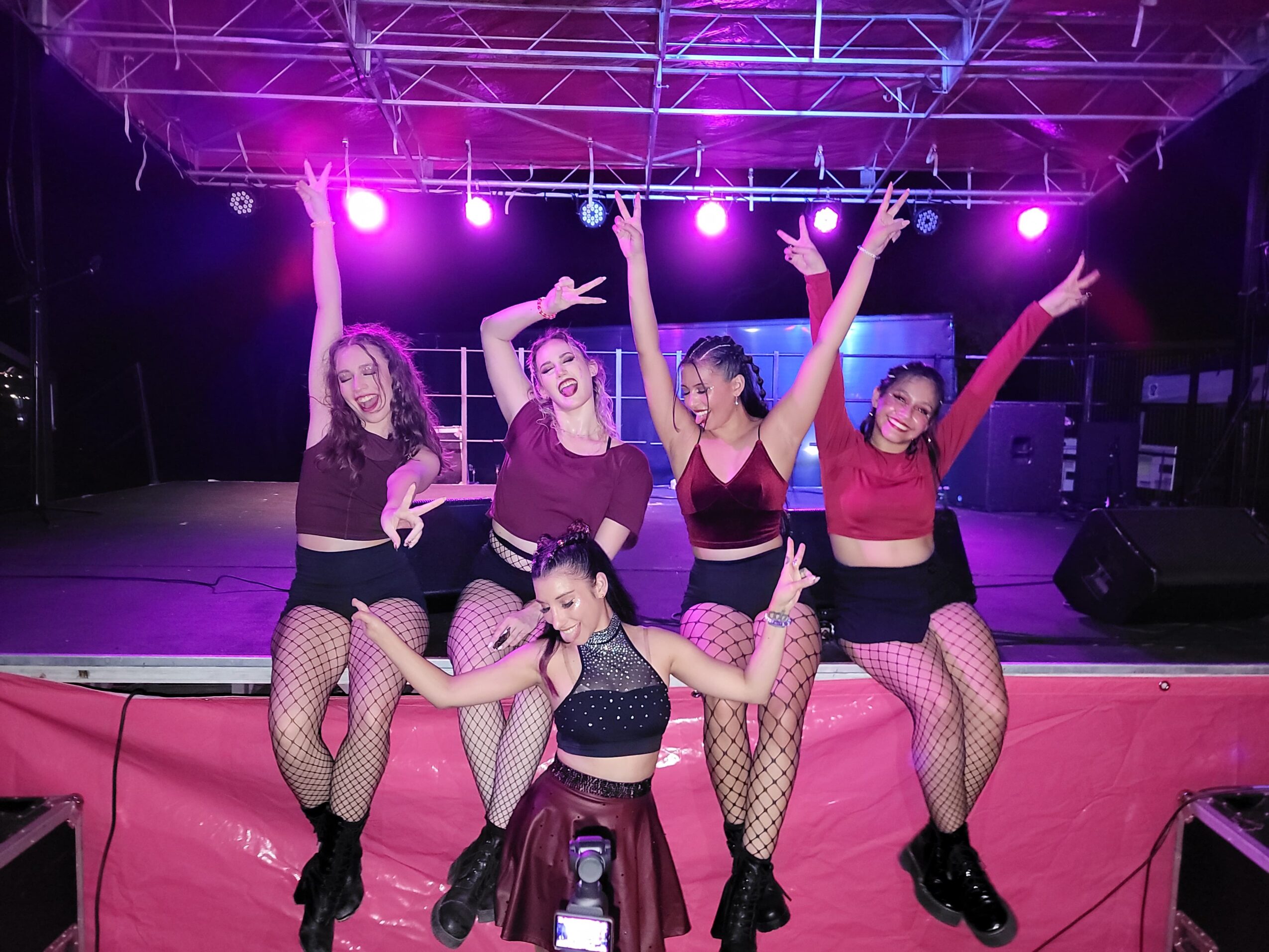 Five young women perform on stage with purple lighting, wearing dance costumes and fishnet stockings, striking energetic poses.