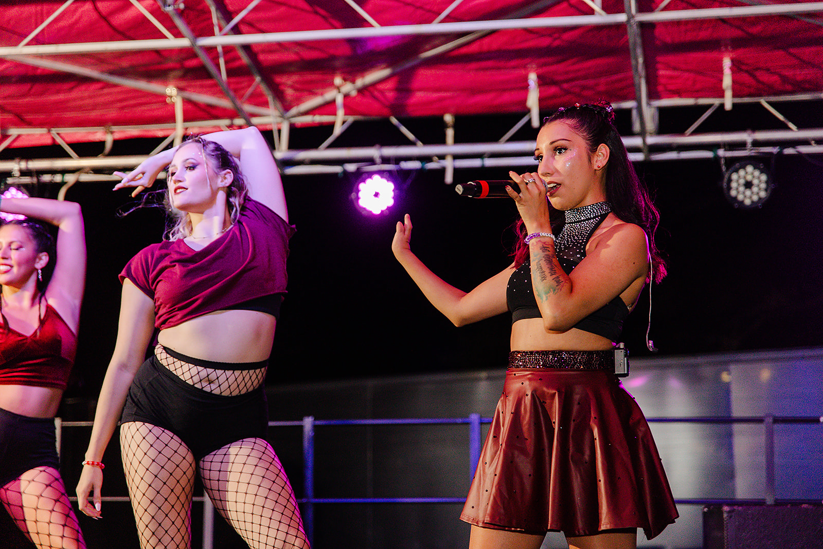Two women performing on stage under a red canopy, one with hands behind head, the other with hand near face, stage lights visible.